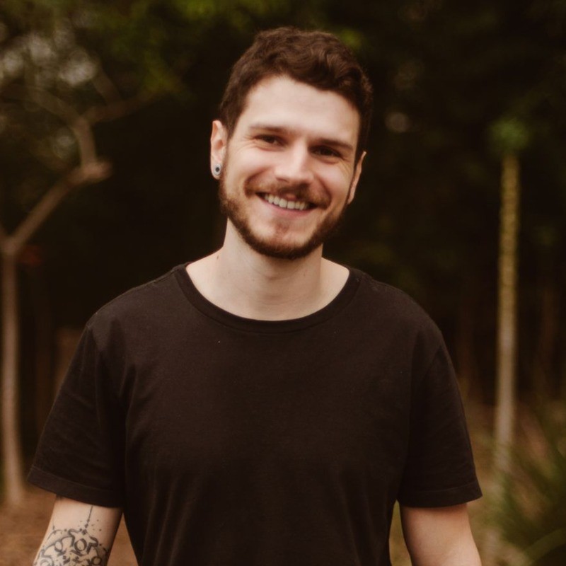 Caio Magro sorrindo, de camiseta preta, em ambiente externo com vegetação ao fundo.